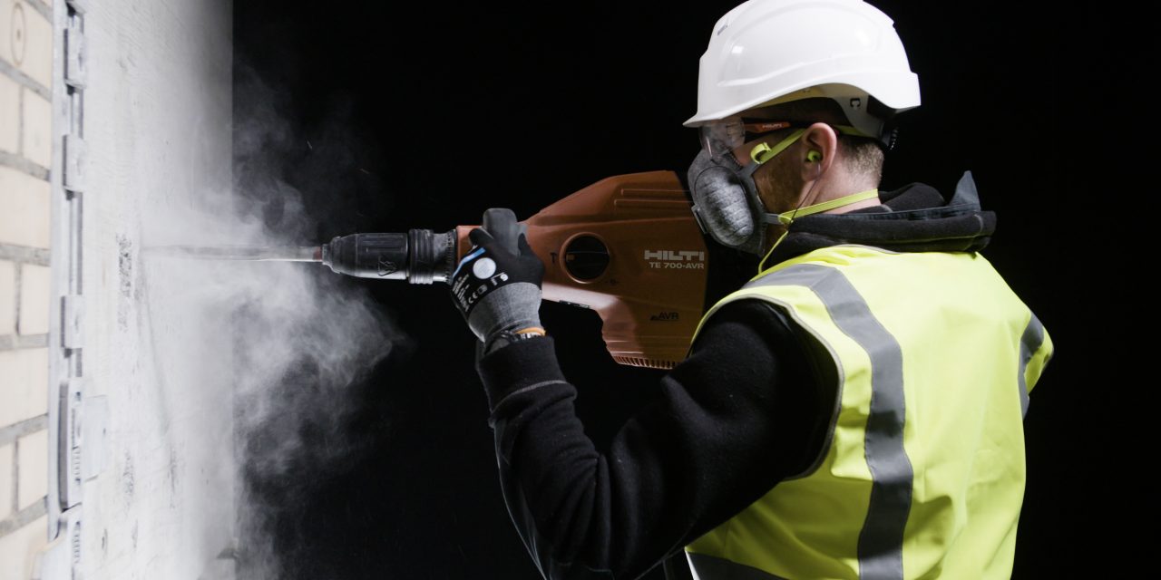 Bauarbeiter mit Atemschutzmaske und Helm arbeitet mit Hilti Bohrhammer an einer Wand und es staubt