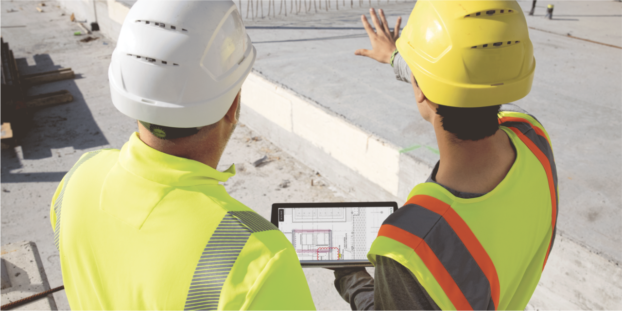 Nahaufnahme von zwei Bauarbeitern, die mit dem Rücken zur Kamera stehen und den Blick auf eine Baustelle gerichtet haben. In ihrer Hand ein Tablet mit digitalen Bauplänen.