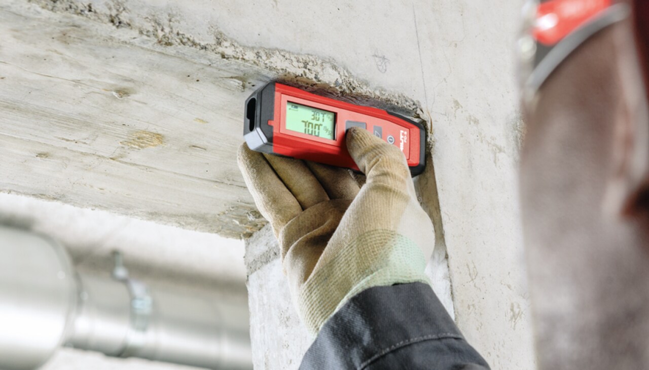 Eine Hand in Arbeitshandschuhen, die mit einem Lasermessgerät Abstände in einem Rohbau prüft.