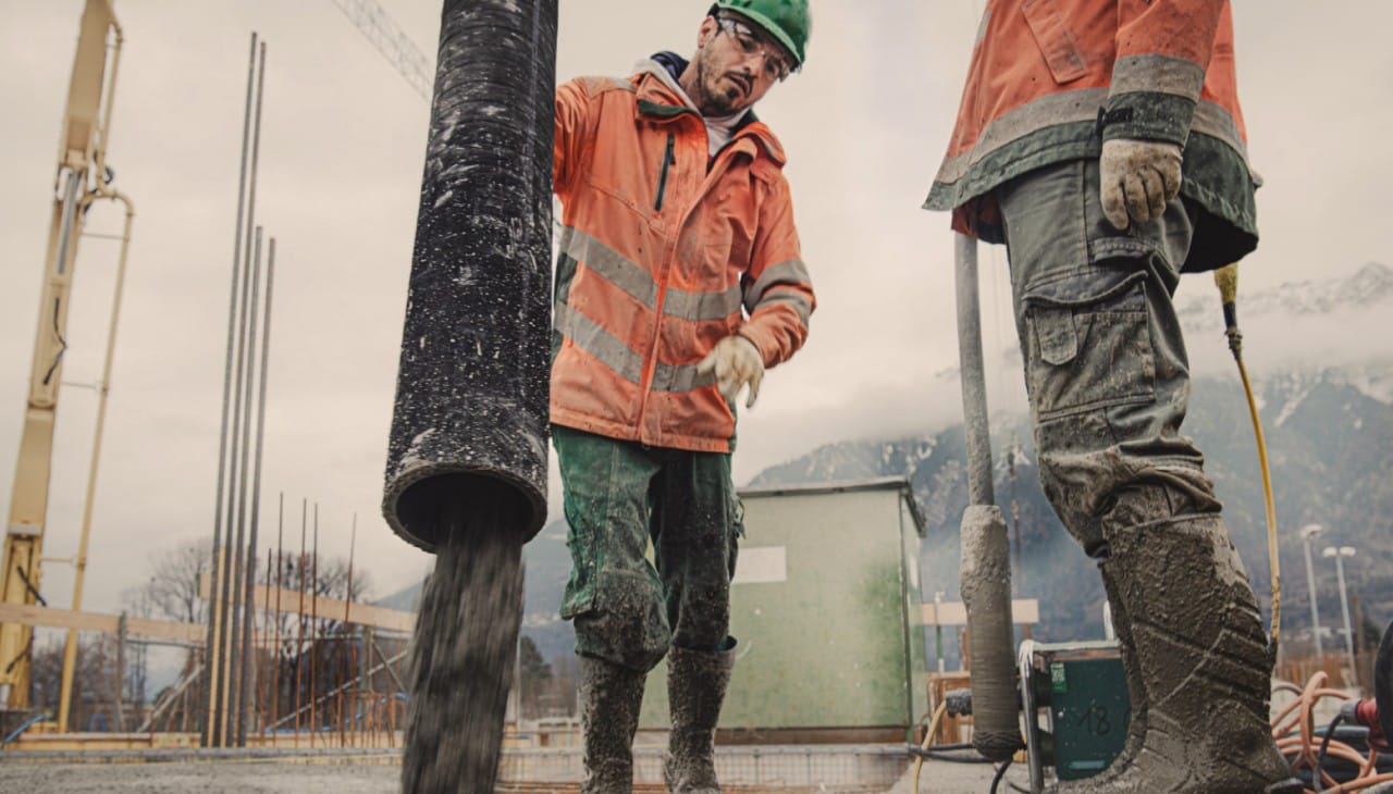 Zwei Männer stehen auf einer Baustelle neben einem Rohr, das eine Betonmischung ausgibt.