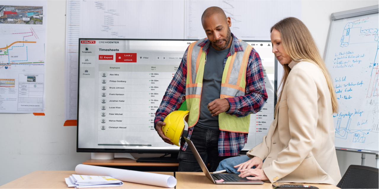 Un homme portant des vêtements de chantier se tient aux côtés d'une femme dans un bureau. Tous deux regardent l’ordinateur portable devant eux.