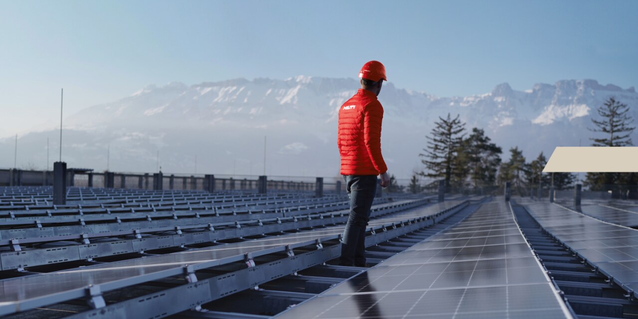 Un homme portant une veste Hilti et un casque rouge se tient entre des panneaux solaires et regarde une chaîne de montagnes derrière lui.