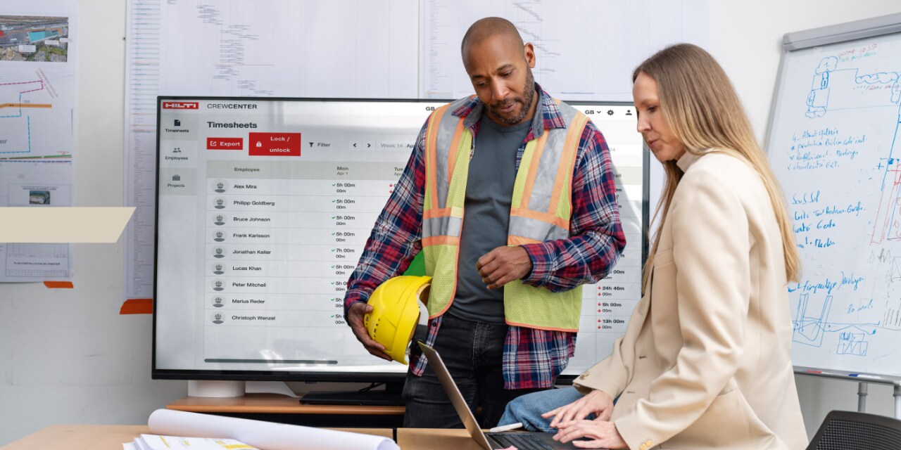 Un homme portant des vêtements de chantier se tient aux côtés d'une femme dans un bureau. Tous deux regardent l’ordinateur portable devant eux.