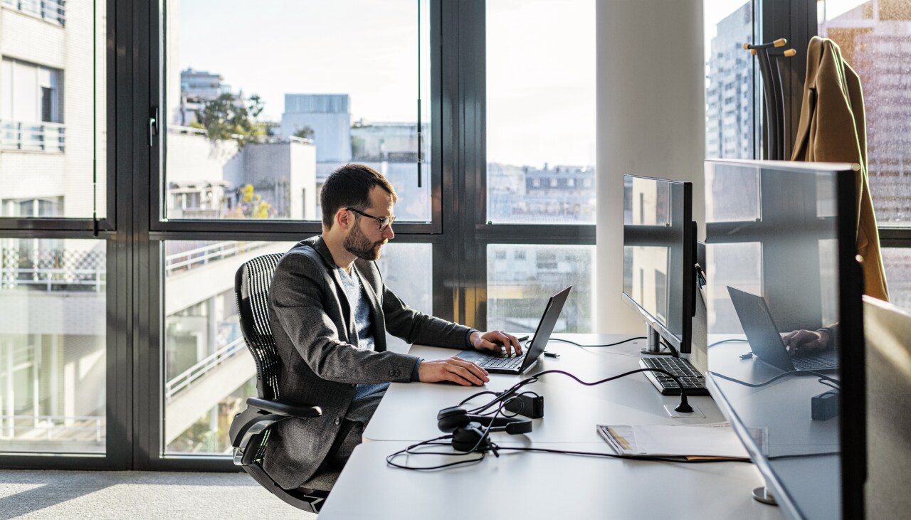 Un homme est assis devant son ordinateur portable dans un bureau avec une façade vitrée.