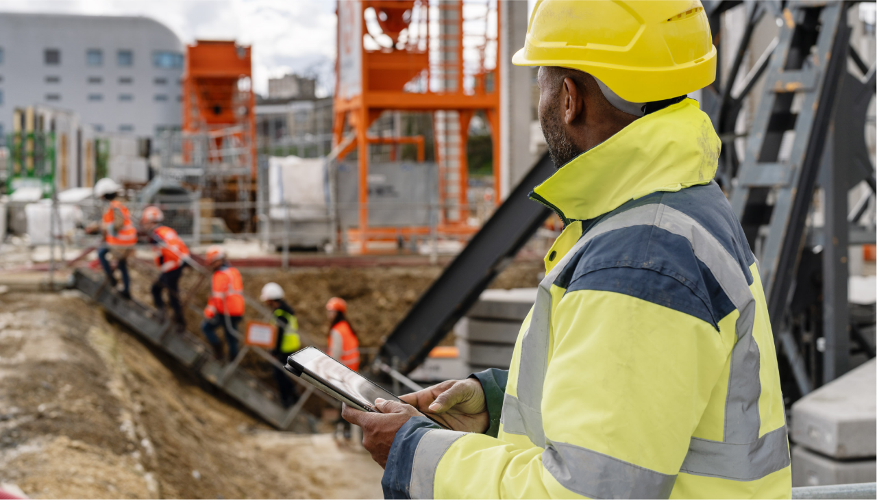 Un uomo che indossa abiti da cantiere e un tablet in mano guarda la fossa di costruzione da cui diversi operai edili stanno salendo una scala.