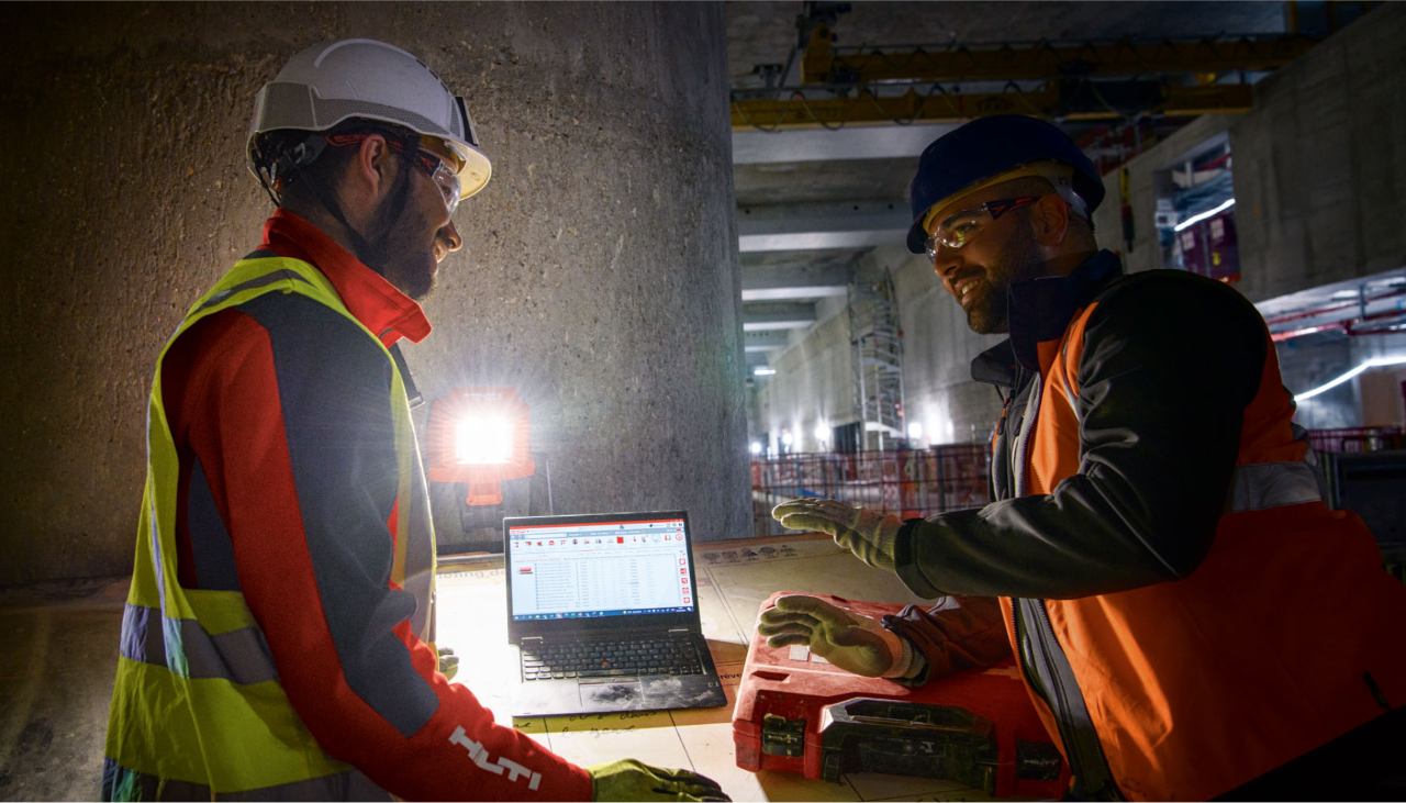 Due uomini vestiti da cantiere parlano tra loro. Di fronte a loro c'è un laptop su un tavolo in un ambiente piuttosto buio.