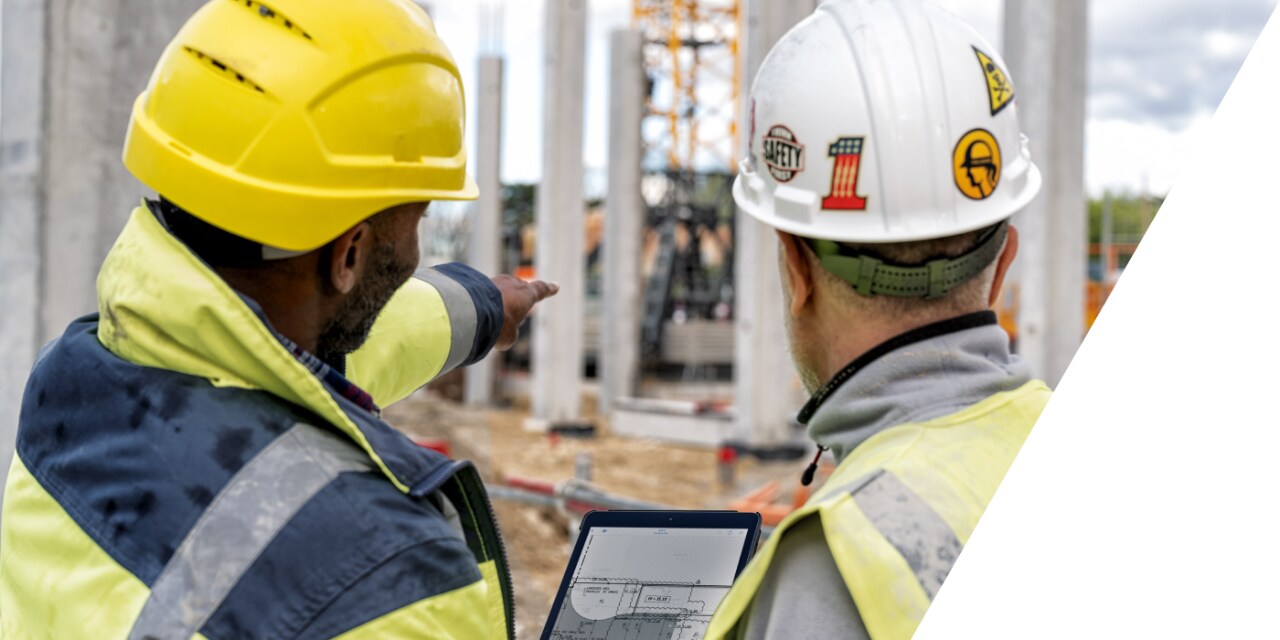 Primo piano di due persone in abiti da cantiere che guardano il cantiere di fronte a loro mentre uno di loro tiene in mano un tablet.